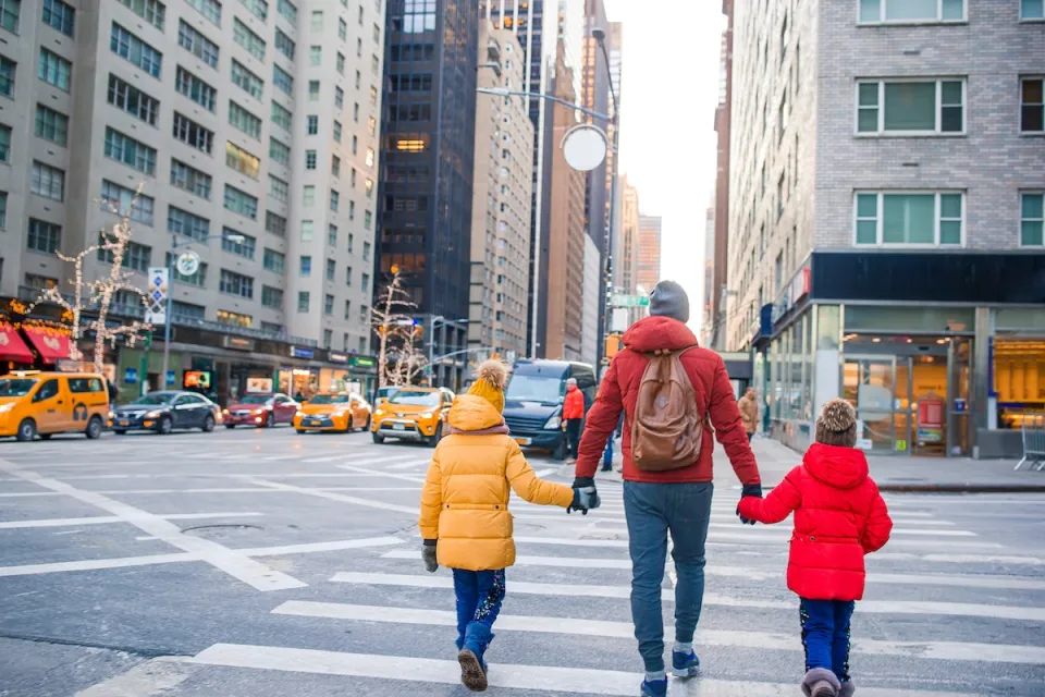 Adult holding hands of two children, all wearing winter coats, in crosswalk in New York City during holidays with trees decorated with lights in background.